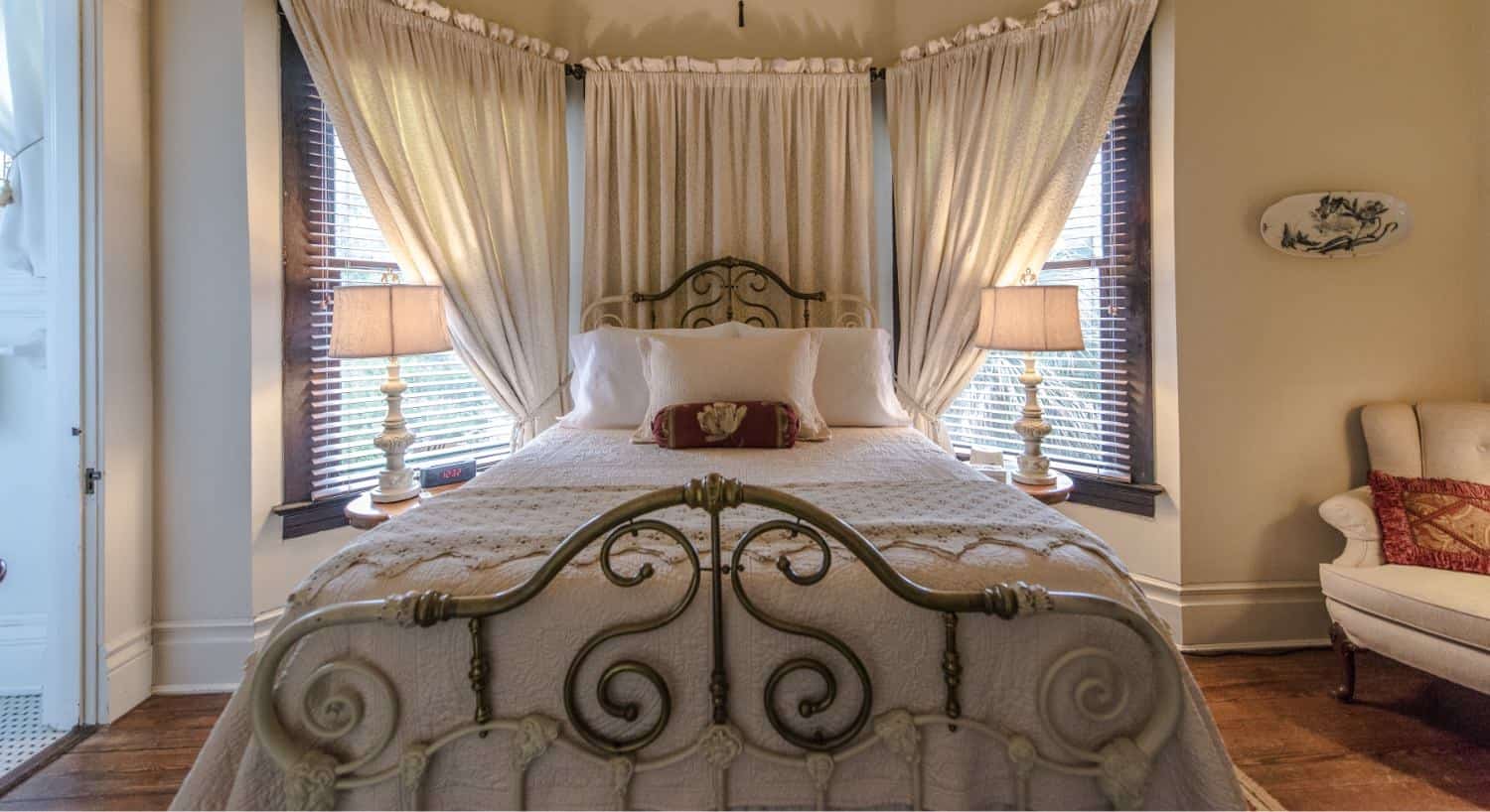 Bedroom with cream-colored walls, hardwood floors, iron bed with cream bedding, and cream upholstered chair