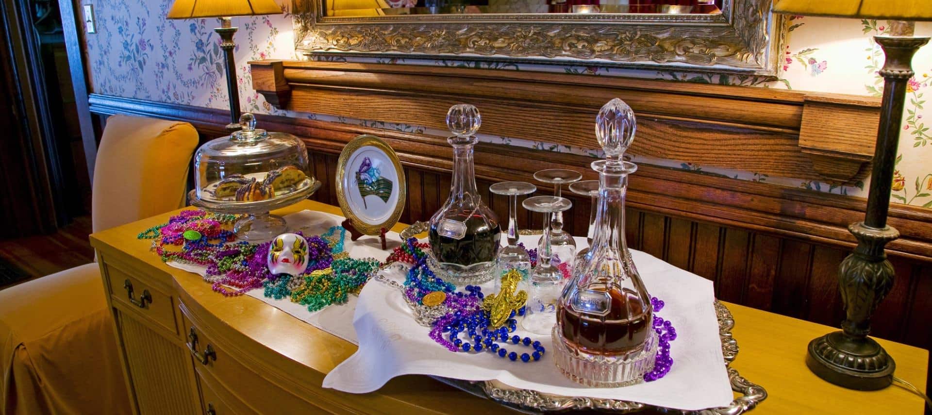 Large wooden buffet topped with Mardi Gras decorations, pastries in glass container, alcohol in glass decanters, and wine glasses